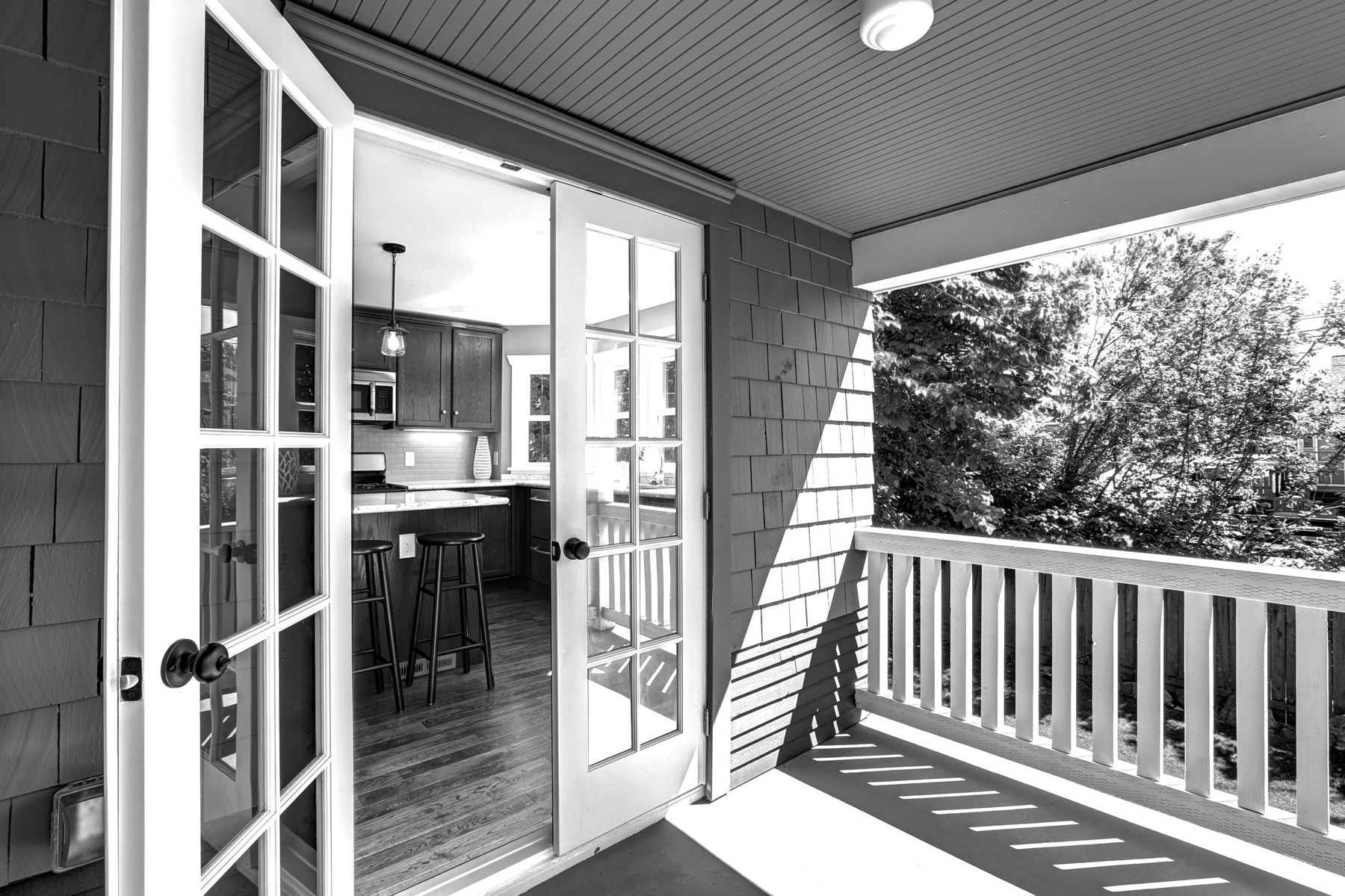 Black-and-white photograph of a covered porch with white railings and open french doors looking into a kitchen.